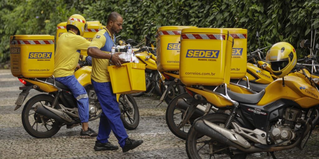 Carteiros uniformizados organizando objetos postais ao lado de motocicletas amarelas com baús do Sedex estacionadas em um pátio de unidade dos Correios.