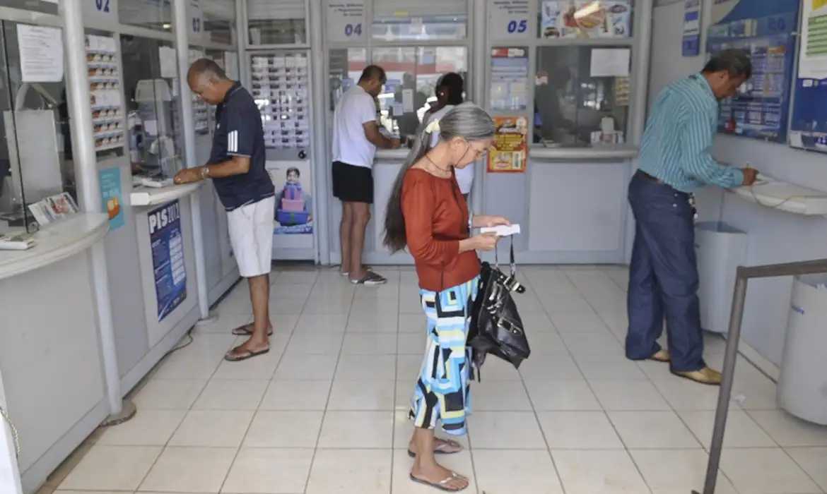 Clientes em uma casa lotérica, alguns nos guichês de atendimento e uma apostadora conferindo o bilhete em pé no salão.