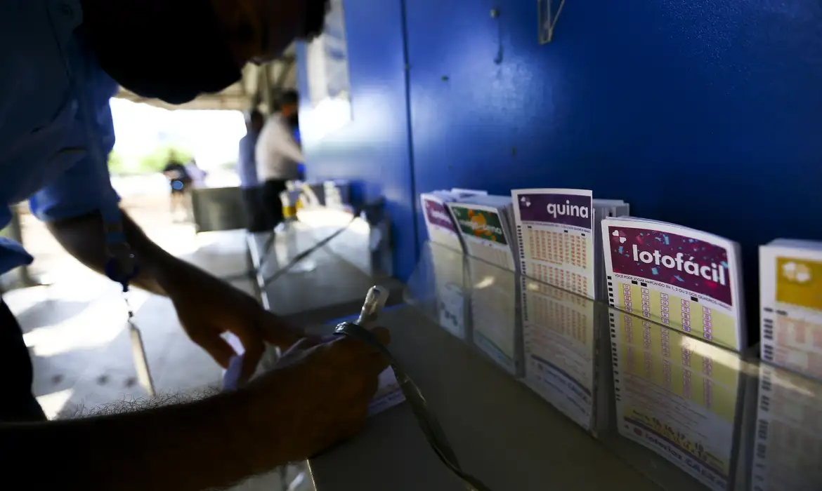 Apostador preenchendo um volante em uma casa lotérica, com cartelas da Lotofácil e de outras modalidades expostas no balcão ao lado.