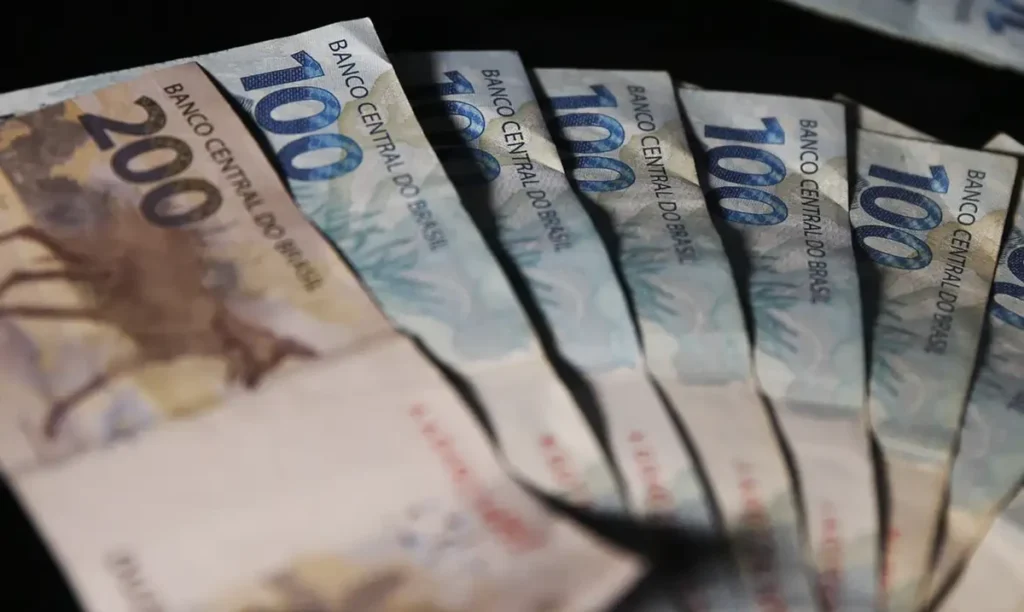 Imagem ilustrativa de cédulas de R$ 200 e R$ 100 dispostas em leque sobre fundo escuro, em referência à consulta do calendário do INSS