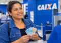 Imagem ilustrativa de mulher sorridente segurando cédulas de R$ 100 e R$ 50 no balcão de atendimento de uma agência da Caixa, em referência ao pagamento do Bolsa Família