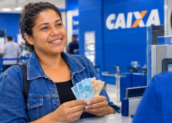 Imagem ilustrativa de mulher sorridente segurando cédulas de R$ 100 e R$ 50 no balcão de atendimento de uma agência da Caixa, em referência ao pagamento do Bolsa Família