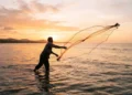 Pescador artesanal lançando rede ao mar durante o pôr do sol, representando pagamento do INSS para trabalhadores da pesca.