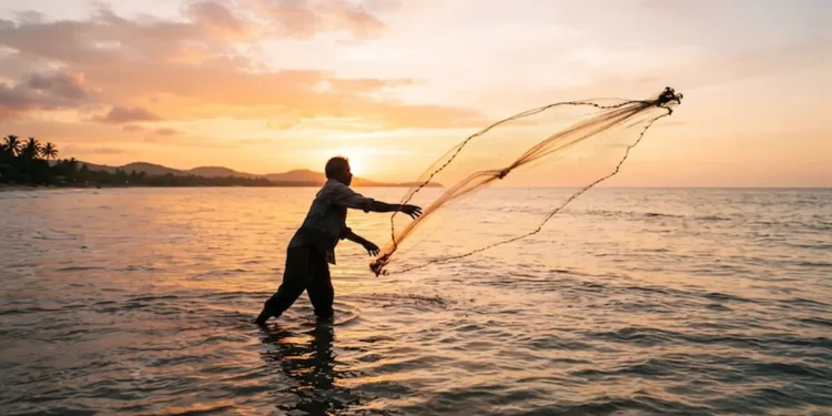 Pescador artesanal lançando rede ao mar durante o pôr do sol, representando pagamento do INSS para trabalhadores da pesca.