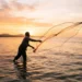 Pescador artesanal lançando rede ao mar durante o pôr do sol, representando pagamento do INSS para trabalhadores da pesca.