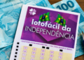 Volante da Lotofácil da Independência apoiado sobre notas de cem reais, com caneta ao lado, em cima de uma mesa de madeira.