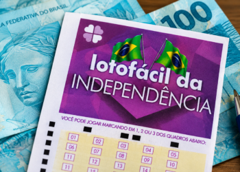 Volante da Lotofácil da Independência apoiado sobre notas de cem reais, com caneta ao lado, em cima de uma mesa de madeira.