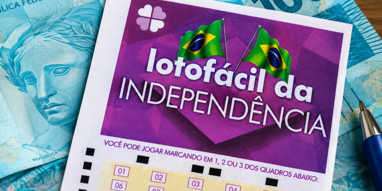 Volante da Lotofácil da Independência apoiado sobre notas de cem reais, com caneta ao lado, em cima de uma mesa de madeira.