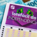 Volante da Lotofácil da Independência apoiado sobre notas de cem reais, com caneta ao lado, em cima de uma mesa de madeira.