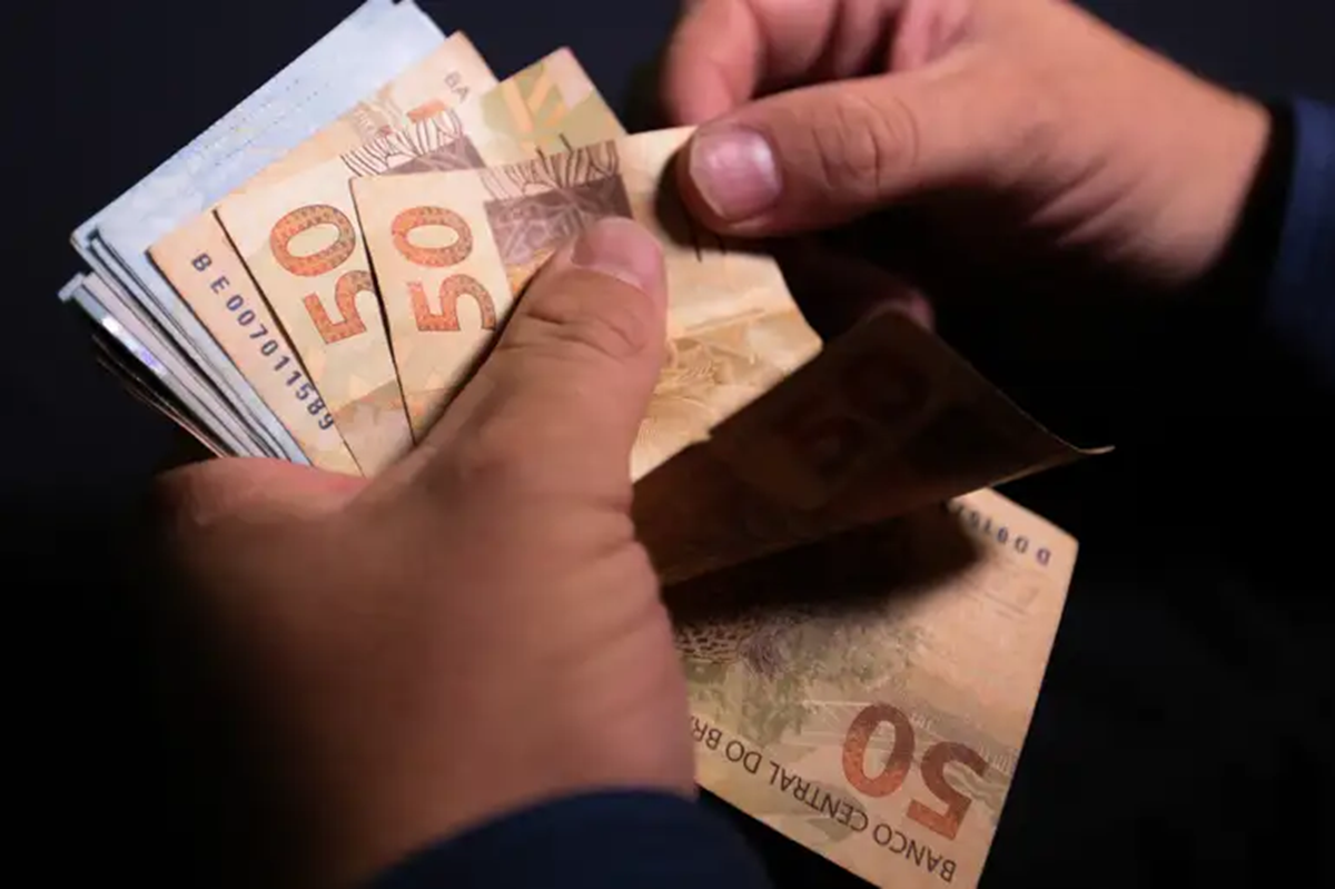 Imagem ilustrativa: mãos contando cédulas de 50 reais, cena que remete ao custo do reajuste no Bolsa Família