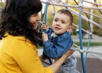 Imagem ilustrativa de mãe segurando o filho pequeno em uma estrutura de cordas de um parque infantil ao ar livre.