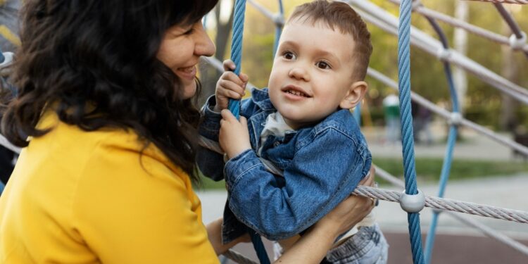Imagem ilustrativa de mãe segurando o filho pequeno em uma estrutura de cordas de um parque infantil ao ar livre.