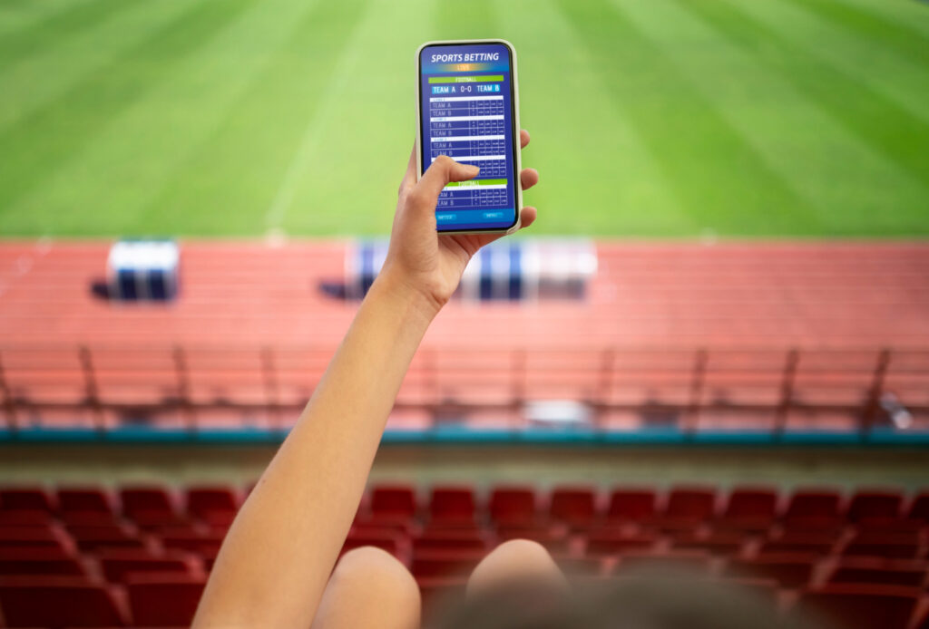 Imagem ilustrativa de mão segurando um celular com uma tela de apostas esportivas aberta, tendo ao fundo o gramado e as arquibancadas de um estádio.