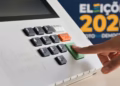 Imagem ilustrativa de dedo pressionando a tecla Confirma da urna eletrônica, com painel das Eleições 2026 ao fundo, em referência às regras do dia da votação