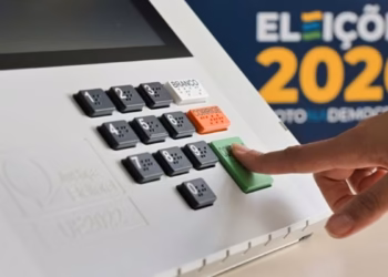 Imagem ilustrativa de dedo pressionando a tecla Confirma da urna eletrônica, com painel das Eleições 2026 ao fundo, em referência às regras do dia da votação